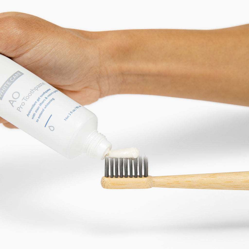 Hand squeezing AO Pro Post-Whitening Toothpaste from white tube onto a bamboo toothbrush with charcoal bristles